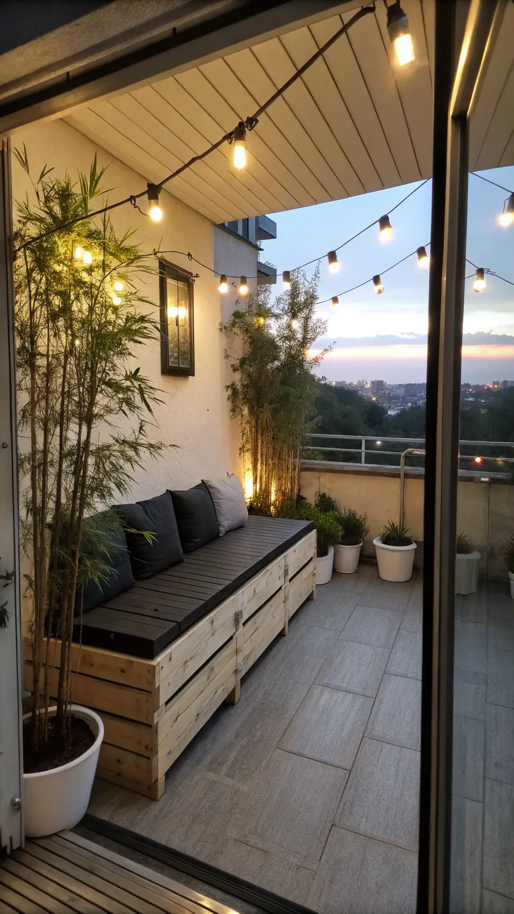 Twilight view of a contemporary 5x9ft balcony with bench seating made from black-stained pallets, bamboo in white ceramic planters, industrial string lights overhead, and brushed steel side tables with lanterns.