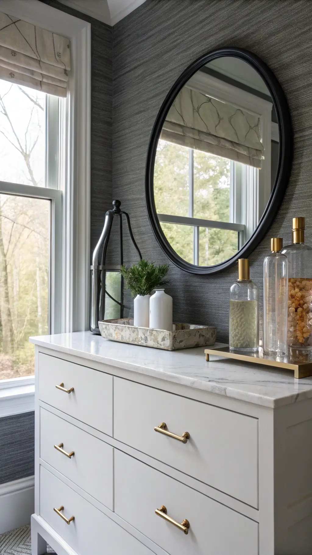 Elegant window-side vanity with white marble top and brass hardware, surrounded by glass and ceramic decor, set against charcoal grasscloth wallpaper, all bathed in cool morning light reflected by oversize round mirror.