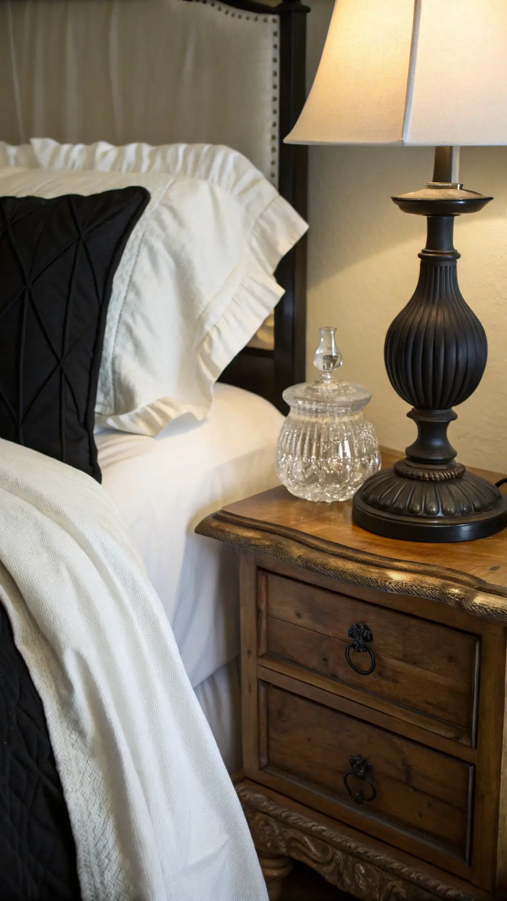 Vintage black table lamp on a rough-hewn wooden nightstand next to a bed draped in white linen and black silk bedding in evening light, with crystal and matte black objects reflecting softly