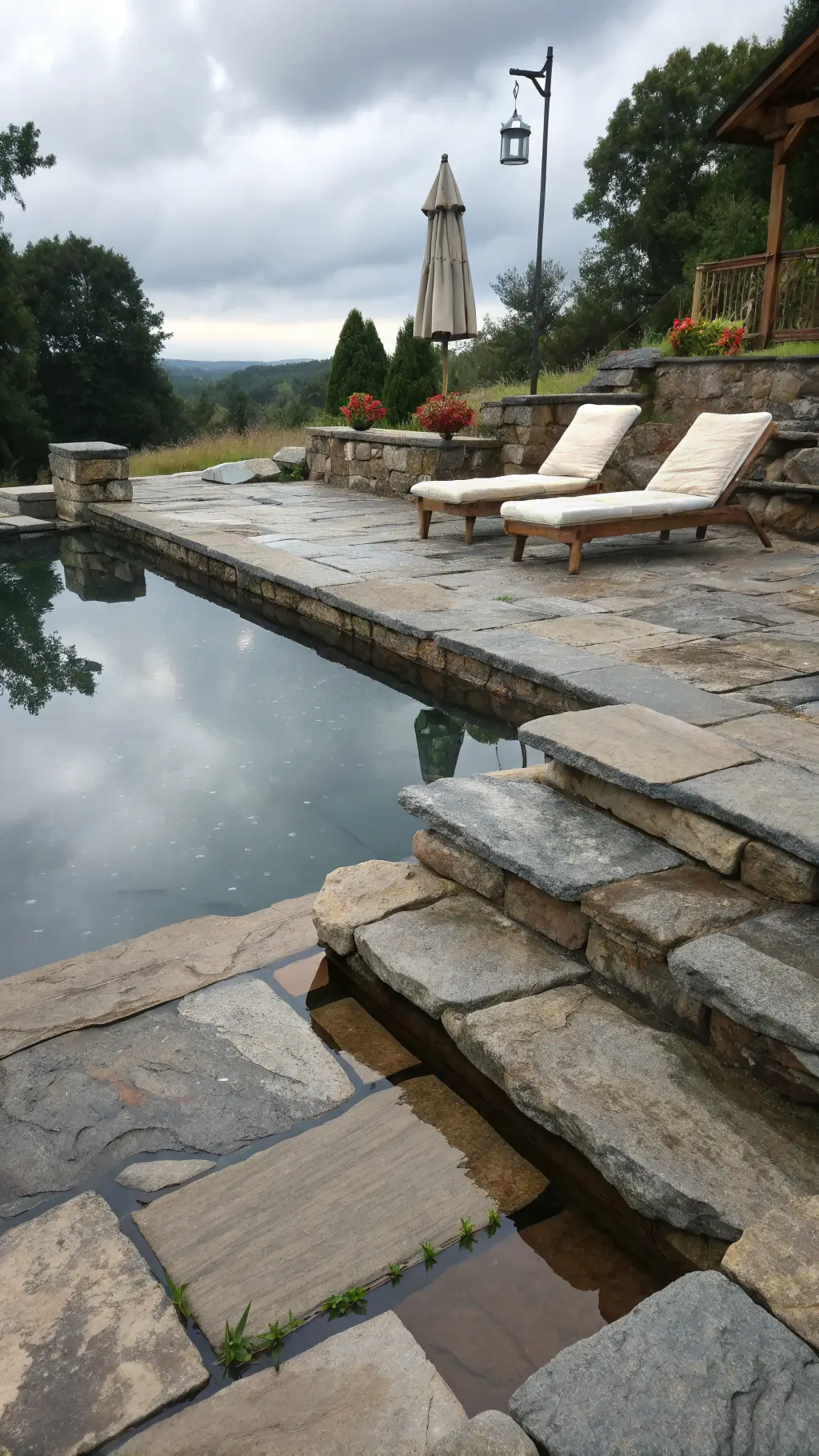 Overcast day illuminating a natural poolside setting with ancient stone steps, weathered wooden loungers, and copper rain chains with a monochromatic gray palette and blue undertones.