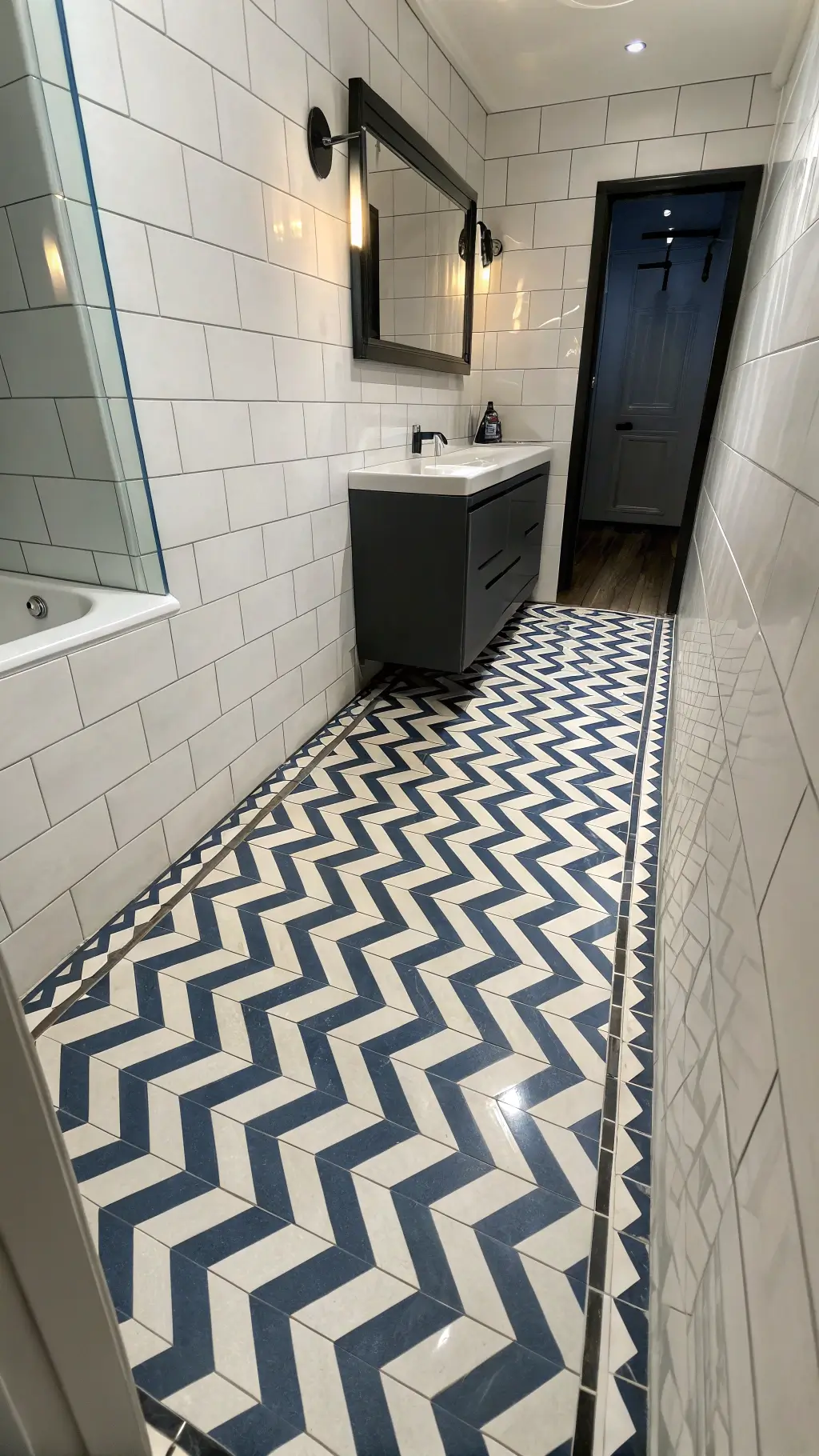 Aerial view of a narrow, sophisticated bathroom with navy blue and white chevron floor tiles, large-format white wall tiles with subtle veining, and a floating matte black vanity under recessed lighting, creating bold and geometric directional flow.